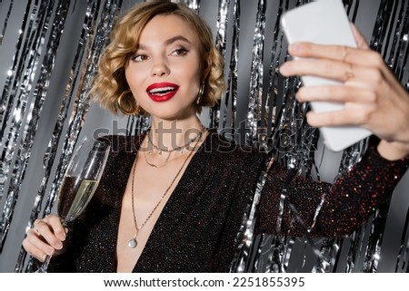 cheerful woman in black dress taking selfie while holding glass of champagne near tinsel curtain on grey Royalty-Free Stock Photo #2251855395