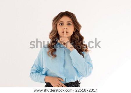 Shush, be silent! Serious lovely female asks to keep secret information confidential, dressed in blue oversized shirt, poses against white studio wall. Conspiracy concept