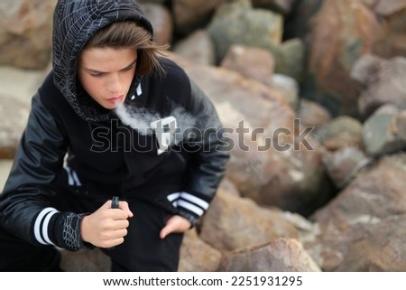 a guy of thirteen years old, holds a vape in his hands. A teenager smokes a disposable electronic cigarette while sitting by the sea Royalty-Free Stock Photo #2251931295