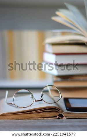 Stack of books on the table, reading glasses and e-reader on the table. Bookshelf in the background. Selective focus.