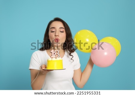 Coming of age party - 18th birthday. Woman blowing number shaped candles on cake and holding balloons against light blue background