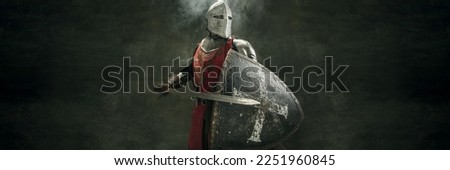 Serious brutal bearded man, medieval warrior and knight with dirty wounded face holding big sword over dark background. Concept of courage, history. Banner with copy space for ad Royalty-Free Stock Photo #2251960845