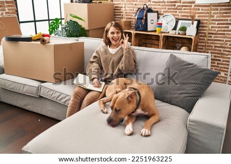 Young beautiful woman sitting on the sofa at new home doing ok sign with fingers, smiling friendly gesturing excellent symbol 