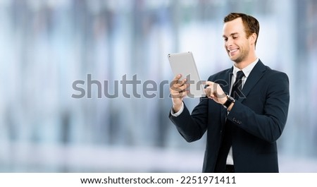Portrait of smiling businessman in black suit using touchpad tablet pc, blurred interior background. Success in business concept studio picture. Copy space for ad text.