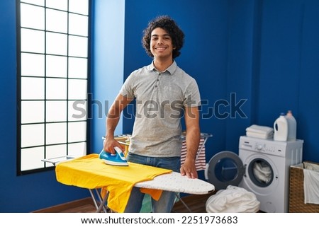Young hispanic man smiling confident ironing clothes at laundry room