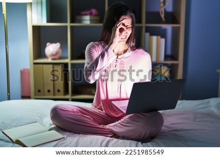 Young hispanic woman using computer laptop on the bed doing ok gesture with hand smiling, eye looking through fingers with happy face. 