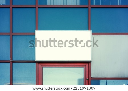 White blank mockup for commercial information in steel frame fixed on glass building wall outdoor over office or store entrance.