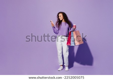 Full body young woman wear pullover hold in hand paper package bags after shopping cup coffee use mobile cell phone isolated on plain pastel light purple background. Black Friday sale buy day concept