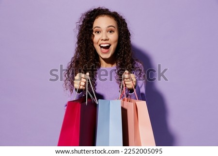 Young surprised woman wear pullover hold in hand paper package bags after shopping look at purchases isolated on plain pastel light purple color background studio. Black Friday sale buy day concept