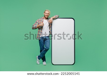Full body young blond man with dreadlocks wear casual shirt big huge blank screen mobile cell phone with workspace copy space mockup area show thumb up isolated on pastel plain light green background