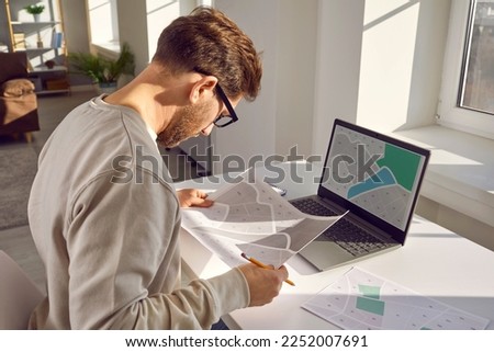 Side view portrait of a professional cartographer working with printed cadastral map at table on his workplace. Young man analyzing cadastral map on laptop and searching for a building plot. Royalty-Free Stock Photo #2252007691