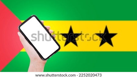 Close-up of male hand holding smartphone with blank on screen, on background of blurred flag of Sao Tome and Principe.