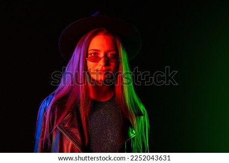 Fashionable woman in neon multi-color light. Smoky background, night party, disco club. Z-generation enigmatic beautiful lady in leather jacket, hat, eyewear. High quality photo