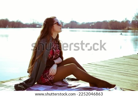 A young woman sits on a wooden pier on a plaid and poses.A woman with long hair in sunglasses.The girl is sitting near the lake.Cold weather.The girl is sitting in a beautiful pose.Vintage and retro.