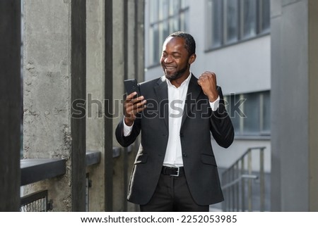 Successful african american boss outside office building using phone businessman in suit celebrating victory, successful achievement, man holding hand up triumph gesture, reading online notification Royalty-Free Stock Photo #2252053985