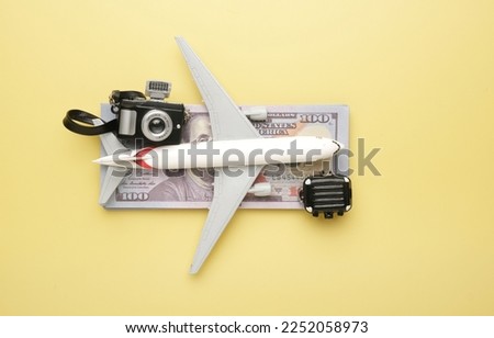 Flatlay picture of toy aeroplane, toy camera, luggage with fake money on yellow background. Expensive flight fare.