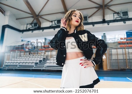 A cheerleader wearing black and white uniform with a black jumper posing in a sports hall. The stands are blurred in the background. High quality photo