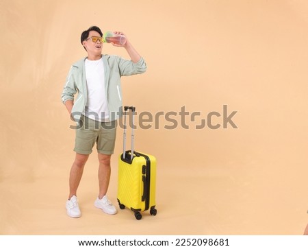 Full body length of a young handsome happy cheerful Asian male adult man, with yellow luggage, ready for travel, isolated on background. Concept of enjoy the trip with good energy drink beverage.