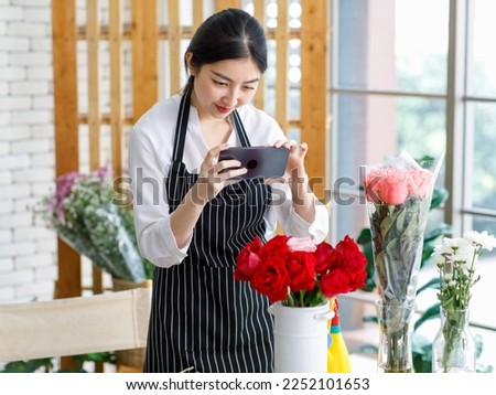 Asian young female flower shopkeeper decorator florist employee worker in apron standing smiling using smartphone taking photo red pink roses bunch bouquet in white ceramic vase on workshop table.