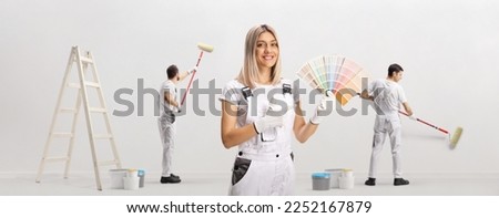 Female house painter holding a color swatch palette and pointing and other workers painting a wall 