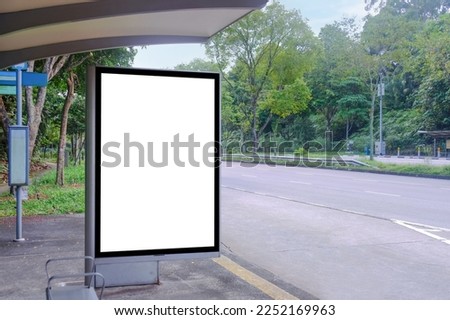 Blank advertising poster banner mockup template at empty bus stop shelter by main road. Greenery around vertical out-of-home OOH billboard advertising display space