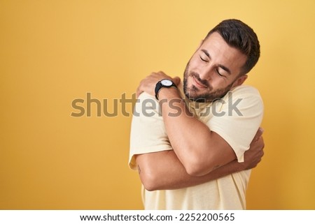 Handsome hispanic man standing over yellow background hugging oneself happy and positive, smiling confident. self love and self care  Royalty-Free Stock Photo #2252200565