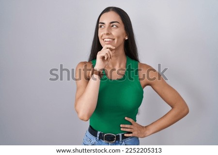 Young woman standing over isolated background with hand on chin thinking about question, pensive expression. smiling and thoughtful face. doubt concept. 