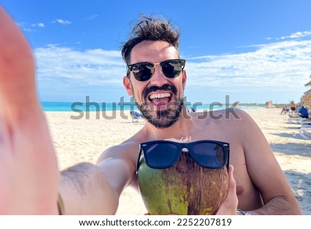 A man enjoys his luxurious vacation in a Maldives resort as he takes a selfie with a coconut and sunglasses. The stunning tropical background sets the perfect scene for a relaxing and carefree holiday