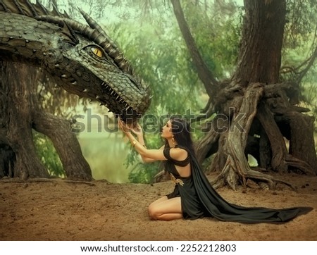 Fantasy woman elf queen touching with hands dragon head. Girl mistress tamed monster concept female power. black creative dress, girl princess fashion model sits on knees. Deep green forest trees.