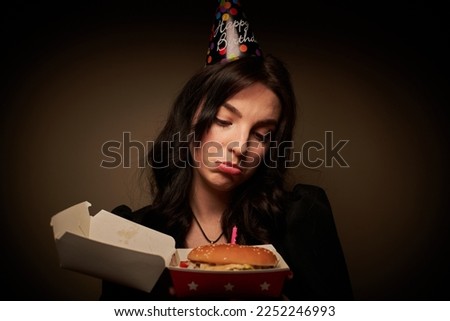 Beautiful brunette girl in a festive hat eats a burger against a brown wall on her birthday instead of a cake  with a candle on a hamburger