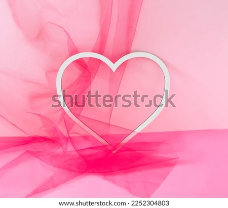 A white frame ih the shape of a heart on pink and magenta background half covered with magenta tulle tissue. Minimal concept for Valentine or wedding card or banner. Copy space.