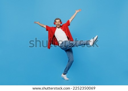 Full body young man of African American ethnicity 20s he wear red shirt headphones listen music dance raise up hands leg isolated on plain pastel light blue cyan background. People lifestyle concept