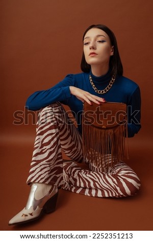 Fashionable confident woman wearing stylish blue turtleneck, trendy flared trousers with zebra print, white cowboy boots, holding brown suede fringed bag. Studio portrait  Royalty-Free Stock Photo #2252351213