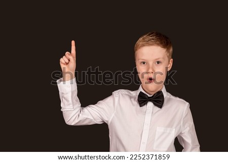 Schoolboy in white shirt with bow tie holding index finger up looking at camera on black isolated background. Good idea, eureka. Stylish little boy student posing in studio. Copy text space for ad