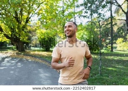 Cheerful and successful hispanic man jogging in the park, man running on a sunny day, smiling and happy having an outdoor activity. Royalty-Free Stock Photo #2252402817