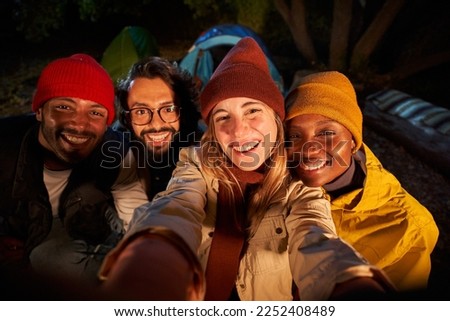 Group of cheerful happy friends outdoors. Photo of men and women smiling and taking selfie in camping forest. Young people with a traveling spirit spending time in the countryside at night. 
