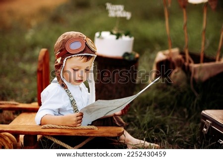 Cute little pilot boy in vintage aviator hat sitting and studying a map near wooden plane outdoor. Happy childhood. Dreams come true.  Royalty-Free Stock Photo #2252430459
