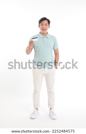 Full body length photo of a standing cheerful young handsome man, holding a credit card, paying online for the bills, isolated on white background
