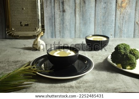 Selective focus picture of Broccoli Soup in a black bowl with broccoli on plate with background blur