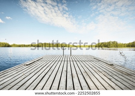Wooden pier near the river (lake). Evergreen trees in the background. Clear blue sky, reflections on the water. Idyllic summer landscape. Ecotourism, recreation, ecological resort, spa, sauna. Finland Royalty-Free Stock Photo #2252502855
