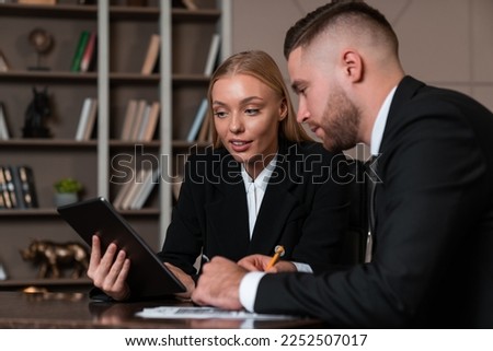 Smiling businessman and businesswoman working together in office room, woman with tablet in hand and man taking notes. Concept of teamwork and communication