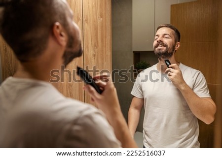 A happy man is holding trimmer and preparing to shave his beard in bathroom. Royalty-Free Stock Photo #2252516037