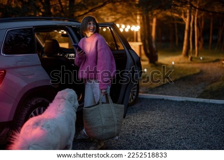 Woman arrives by car to a house in forest, standing with bag and phone near vehicle in the evening time. Traveling by car and rest in cabins on nature concept Royalty-Free Stock Photo #2252518833