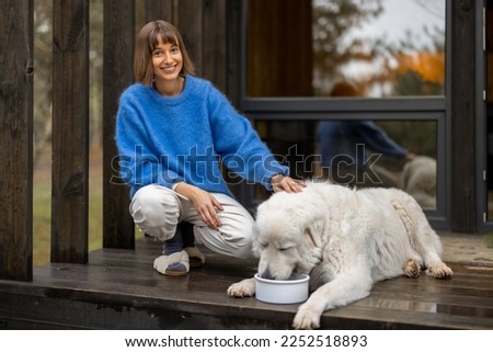 Young woman feeds her cute white dog while sitting together on a porch of wooden house in the forest. Pets caring and rest on nature concept