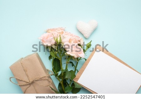 A bouquet of bush roses, a gift in kraft paper, a kraft envelope and a knitted white heart on a light blue background. Valentine's Day, Flat position, top view.