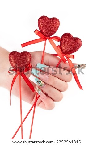 Hand with long artificial manicured nails colored with white and turquoise blue nail polish holding red hearts Valentine day decorations. White background.