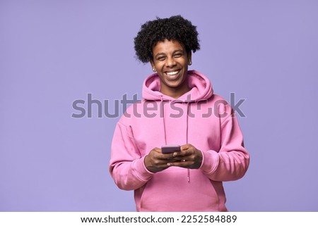 Happy cool curly African American teen guy model wearing pink hoodie holding cell phone using mobile digital apps on cellphone tech texting on smartphone isolated on light purple background.