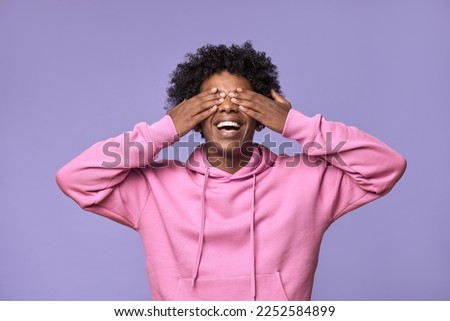 Happy African American teen guy closing eyes with hands feeling amazed waiting for surprise. Smiling blind ethnic student model excited with anticipation standing isolated on light purple background.
