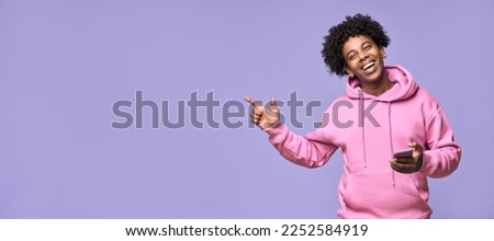 Happy African teenager holding mobile phone pointing finger aside presenting promotion offer, using cellphone showing ads advertising online application standing isolated on light purple background. Royalty-Free Stock Photo #2252584919