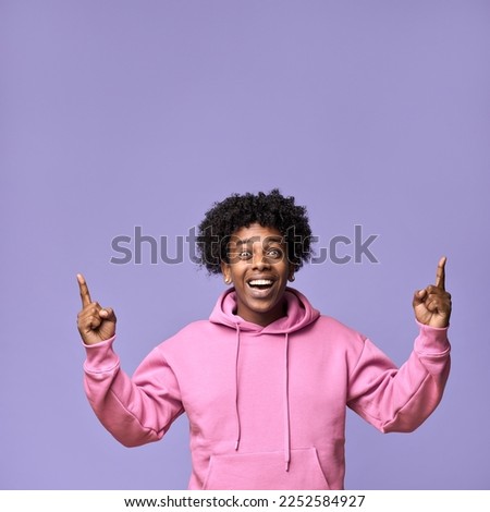 Happy surprised African teen guy looking at camera pointing up advertising sale promotion. Excited ethnic teenager student boy presenting promo offer standing isolated on light purple background.